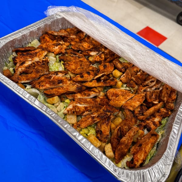 Grilled chicken Caesar salad in a catering tray, partially covered with plastic wrap.