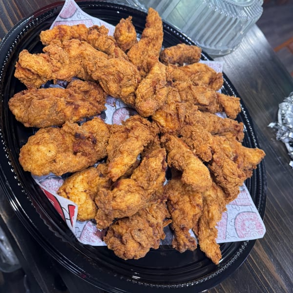 Golden-brown fried chicken tenders piled high on a black catering tray with patterned paper.