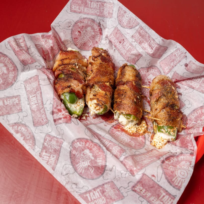 Four seasoned bacon-wrapped jalapeño poppers with creamy cheese, served in a branded basket.