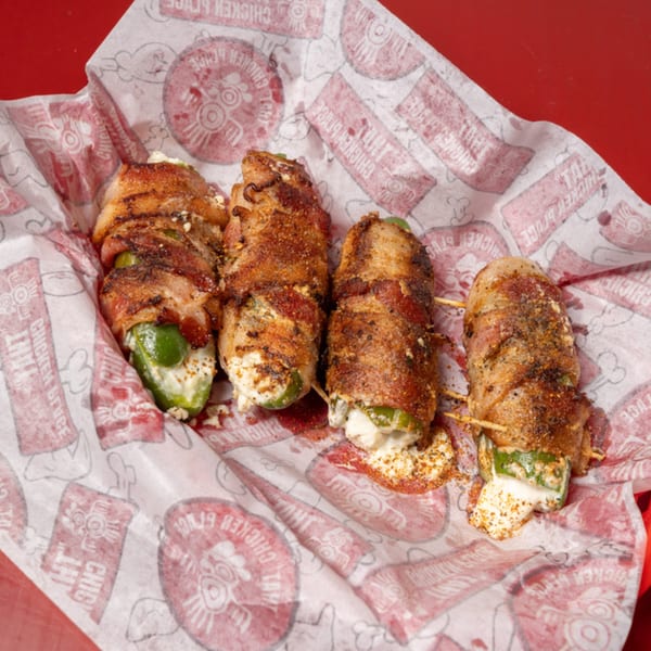 Four crispy bacon-wrapped jalapeño poppers, filled with melted cheese, on branded Chicken Place paper.