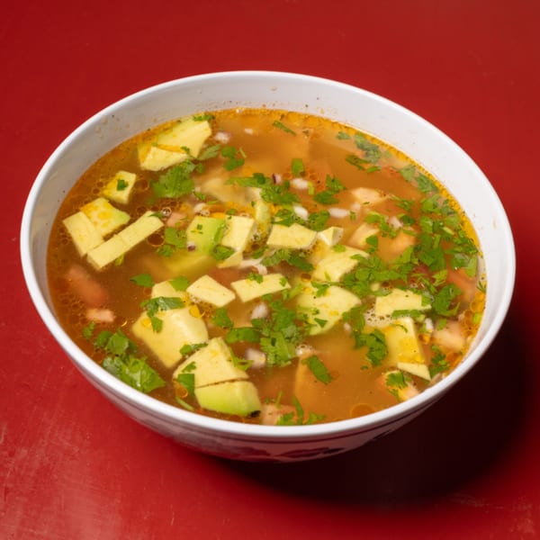 A bowl of chicken soup with avocado, cilantro, and other vegetables on a red background.