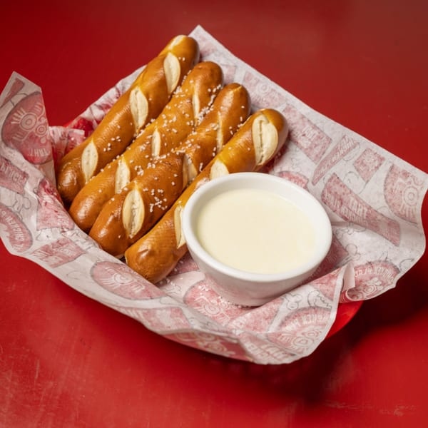 Salted pretzel sticks and a white bowl of cheese dip in a red basket lined with branded paper.