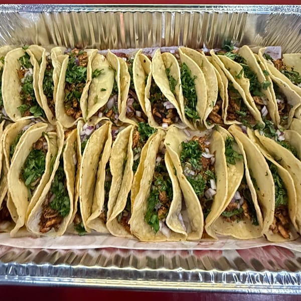 Many tacos with meat, onions, and cilantro in corn tortillas are arranged in a foil catering tray.