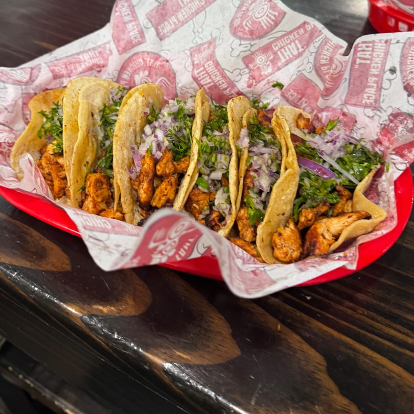 Six chicken tacos topped with red onion and cilantro in a red basket on a wooden table.
